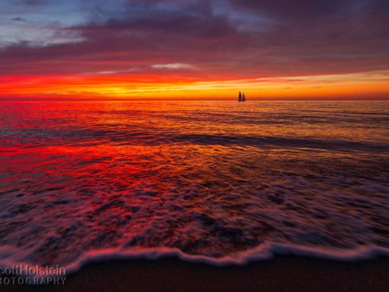 A sailboat sits on the horizon during a vibrant beach sunset in Venice, Florida.