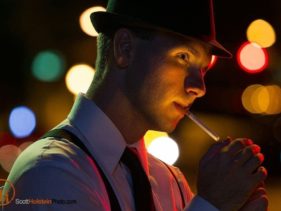 A detective in a fedora lights a cigarette at night in front of glowing street lights in a dramatic portrait.