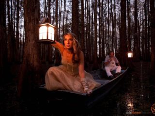 A beautiful woman holding a lantern peers out into a swamp at dusk.
