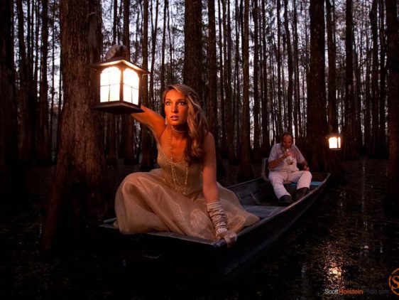 A beautiful woman holding a lantern peers out into a swamp at dusk.